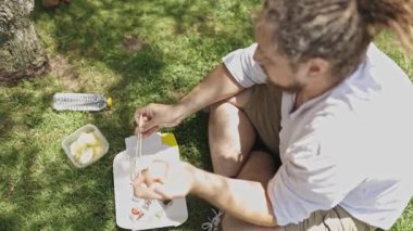 Rastalı bir adamın yüksek açılı görüntüsü. Parktaki çimlerin üzerinde oturmuş suşi yiyor.