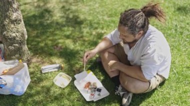 Rasta giymiş rahat bir adam yeşil çimenlerde oturmuş öğle yemeğinde suşi yiyor.