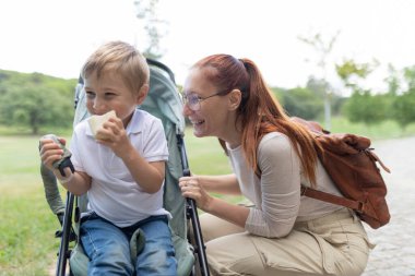 Anne, güneşli bir günde bebek arabasında oturup sandviç yiyen oğlunun yanında çömeliyor.