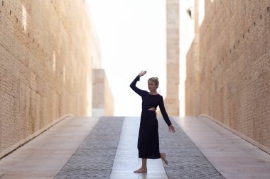 Genç bayan zarif bir şekilde, yüksek mimari duvarlar arasında uzun elbiseler içinde yalınayak hareket ediyor.