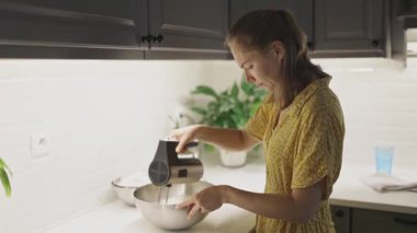 Genç bir kadın, tatlı hazırlarken metal bir kasede elektrikli mikserle krema çırpıyor.
