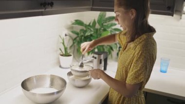 Evde yemek pişirirken metal bir kâseye elek sokup un karıştıran genç bir kadın.