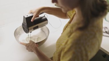 Çelik bir kasede elektrikli el mikseriyle krema çırpan bir kadının yüksek açılı görüntüsü.