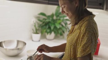 Modern bir mutfakta yemek hazırlayan, kaseye kaşıkla malzeme ekleyen mutlu bir kadın.