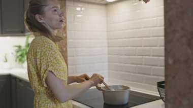 Odaklanmış bir kadın tencerede yemek hazırlıyor, modern bir mutfakta tahta kaşıkla karıştırıyor.