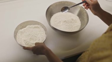 Kadınların elleri, hamur pişirmek için bir kaşıkla süt dolu metal bir kaseye un ekler.