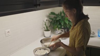 Genç bir kadın beyaz bir tezgahta metal bir kasede un ve sıvı malzemeleri karıştırıyor.