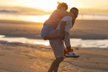 Couple enjoying piggyback ride on sandy beach during golden hour sunset