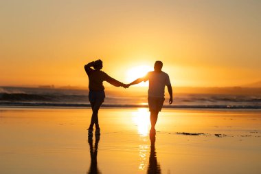 Çiftler gün batımında kumsalda romantik yürüyüşler yaparlar, el ele tutuşurlar ve altın gökyüzüne karşı güzel bir siluet yaratırlar.