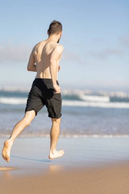 Young man exercising shirtless on a sunny beach, running towards the ocean waves