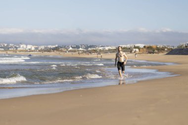 Man running along the waterline on a sunny beach, splashing water while exercising