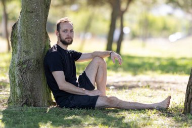 Man enjoying quiet moment sitting barefoot on grass against tree trunk