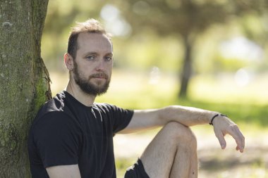 Bearded man sitting in park leaning against tree looking towards the viewer