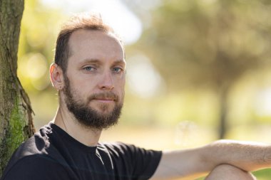Man with beard and blue eyes relaxing outdoors, looking directly at viewer