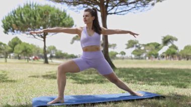 Young woman in sportswear practicing yoga warrior ii pose on a mat in a sunny park
