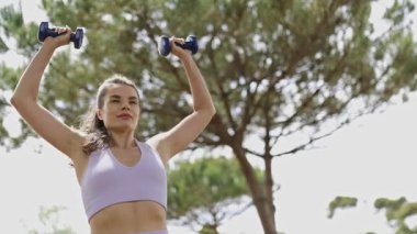 Young athletic woman performing a dumbbell shoulder press exercise outdoors in a park
