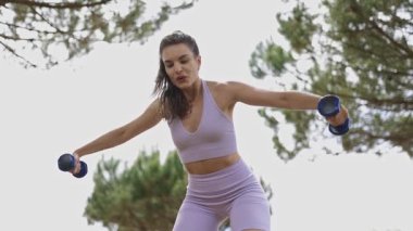 Young athletic woman performing lateral raises with dumbbells during an outdoor workout session