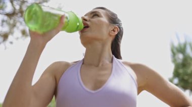 Young athletic woman drinking water from a reusable bottle after a workout outdoors in a park