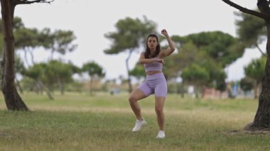 Young woman in sportswear dancing energetically on a grassy lawn in a park with trees