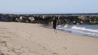 Young man wearing black clothes walking barefoot on a sandy beach along the calm ocean shoreline