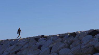Lone man walking on top of a large stone seawall against a clear blue sky