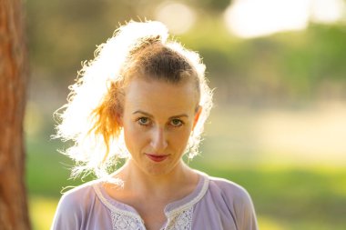 Woman smiling with backlit curly hair in a soft outdoor setting at sunset