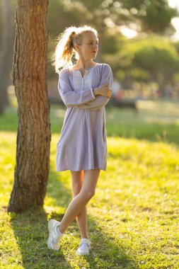 Young woman standing by tree, crossing arms and legs, embracing sunset light in a park
