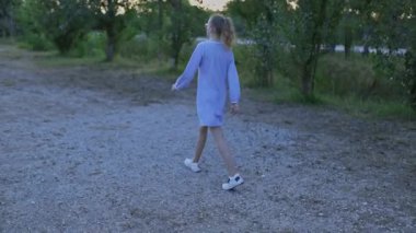 Young girl in a light blue dress is walking on a gravel path in a park at sunset