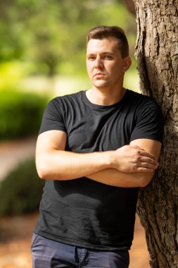 Young man standing with crossed arms, looking thoughtful in an outdoor setting