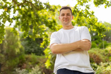Young adult man standing in a park, smiling with his arms crossed on a sunny day