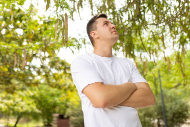 Young man standing with crossed arms outdoors looking up and thinking