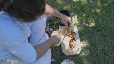 Young man sitting on the grass cutting roasted chicken during a picnic on a summer day