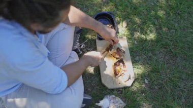 Young man sitting on the grass cutting roasted chicken on a wooden board during a sunny day picnic