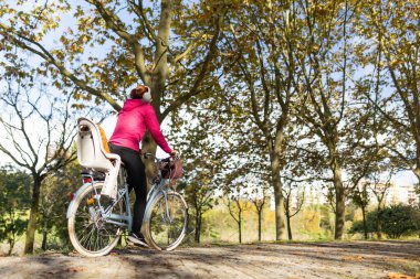 Park yolunda bisiklet süren bir kadın, çocuk koltuğu ile aktif yaşam tarzının tadını çıkarıyor.