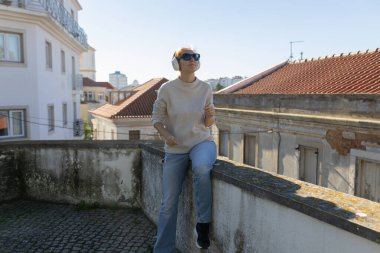 Genç bir kadın, güneşli bir günde duvarda dinleniyor, kulaklıklardan gelen sesten keyif alıyor.