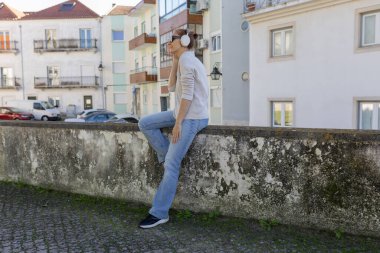 Şehir ortamında alçak bir duvarda oturan bir kadın, kulaklıkla müzik dinlemekten hoşlanıyor.