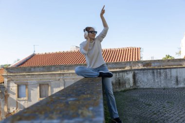 Duvarda oturan, müzik dinleyen ve gökyüzünü neşeyle gösteren genç bir kadın.