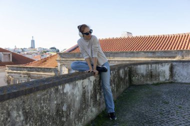 Şehir siluetine tepeden bakan kulaklıklarıyla duvarda oturan rahat bir kadın.