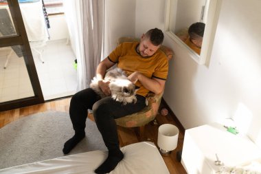 Bir adam güneşli bir odada bir koltukta otururken kediyi tutup okşuyor.