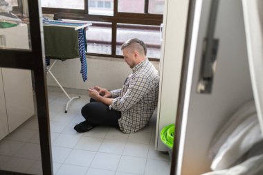 Ev balkonunda fayanslı bir zeminde oturan bir adam cep telefonu kullanıyor.