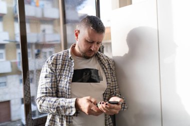 Şehir manzaralı bir pencerenin yanında duran bir adam cep telefonu tutuyor.