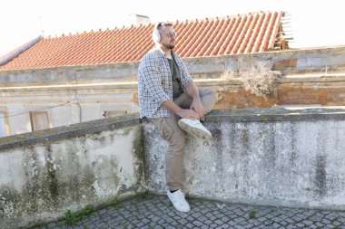 Çatıdaki duvarda oturan, kulaklık takan ve güneş gözlüğü takan genç adam şehir manzarasının tadını çıkarıyor.