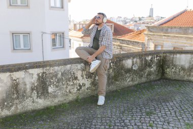 Kulaklıkla müzik dinleyen bir adam şehre tepeden bakan bir duvarda oturuyor.