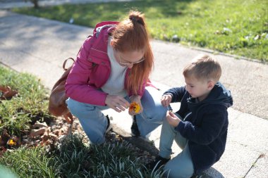 Anne ve oğlu dışarıda çömelmiş, küçük çiçekleri ve bitkileri birlikte inceliyorlar.