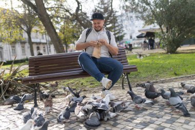 Bankta oturan bir adam şehir parkında kağıt torbadan güvercinleri besliyor.