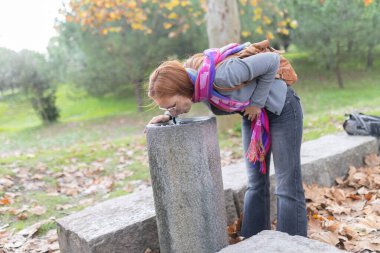 Kadın, sonbahar yapraklarıyla halka açık bir parkta taştan yapılmış bir çeşmeden su içiyor.