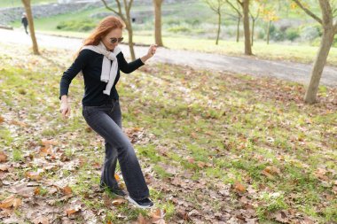 Gülümseyen kadın, sonbahar yaprakları arasında boş zamanlarında parkta dikkatli bir şekilde yürüyor.