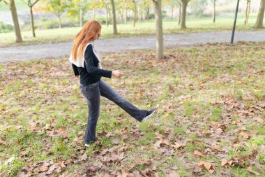 Parkta oynayan kadın, ağaçlarla çevrili çimenlerin üzerinde kuru sonbahar yapraklarını tekmeliyor.