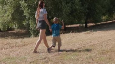 Güneşli bir günde çimenli bir arazide yürürken küçük oğullarının elini tutan genç bir anne.