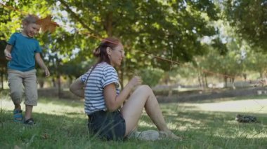 Mutlu bir aile, bir anne ve küçük oğlu, güneşli bir parkta çimlerin üzerinde birlikte gülüp oynuyorlar.